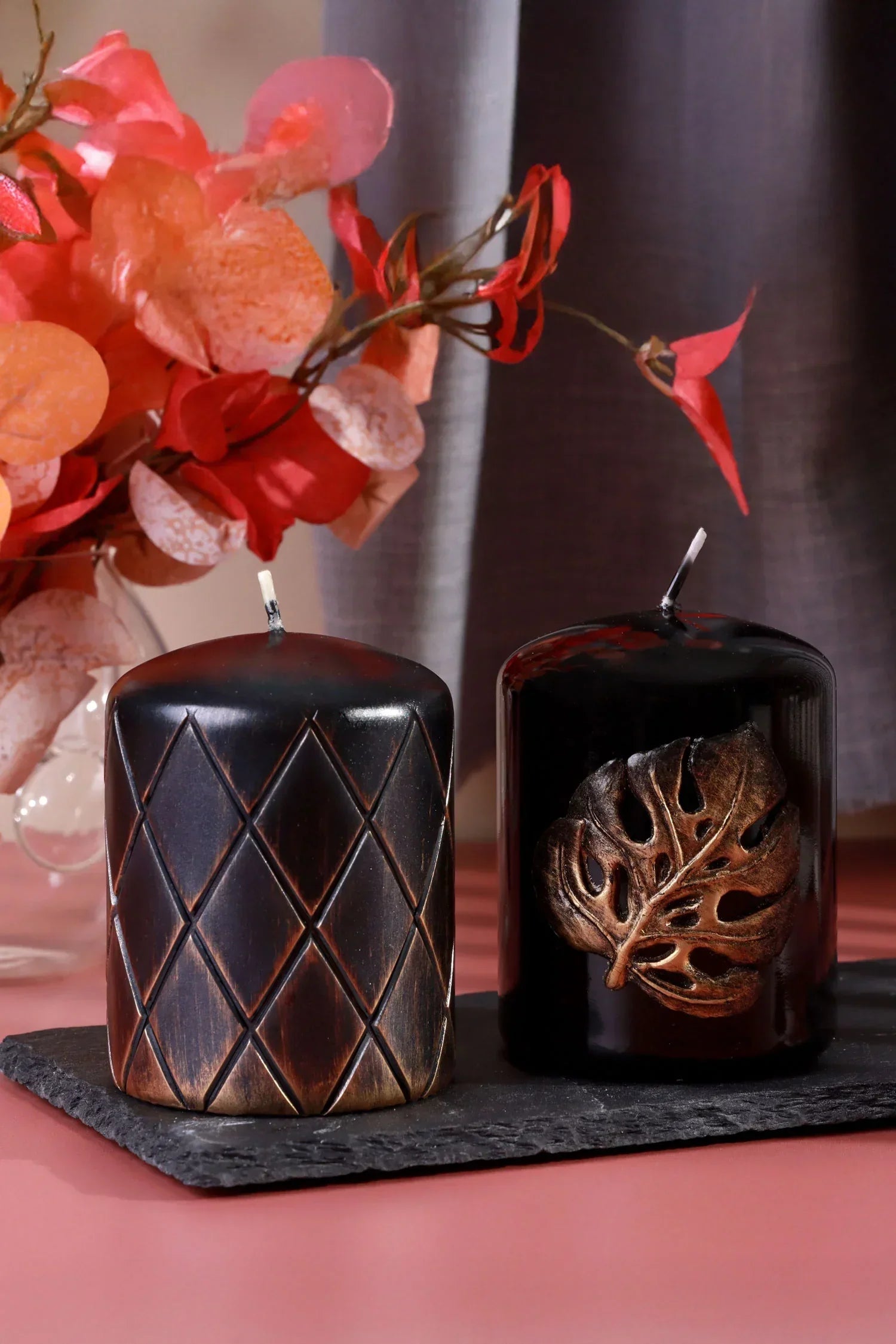 Luxury black pillar candles with geometric and leaf designs on slate, floral decor in background