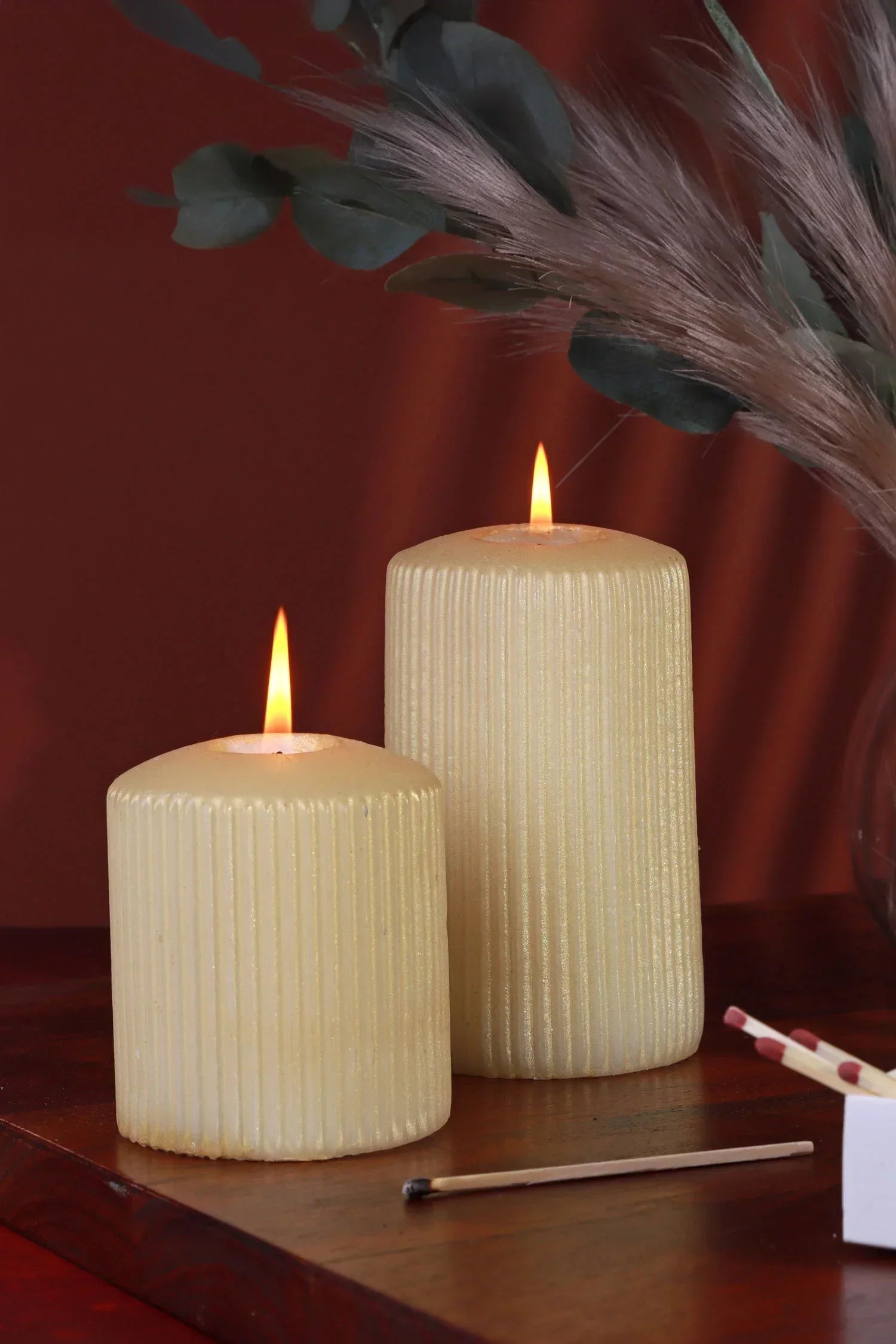 Two ribbed pillar candles burning on a wooden table with dried foliage, luxury home décor by G Decor.