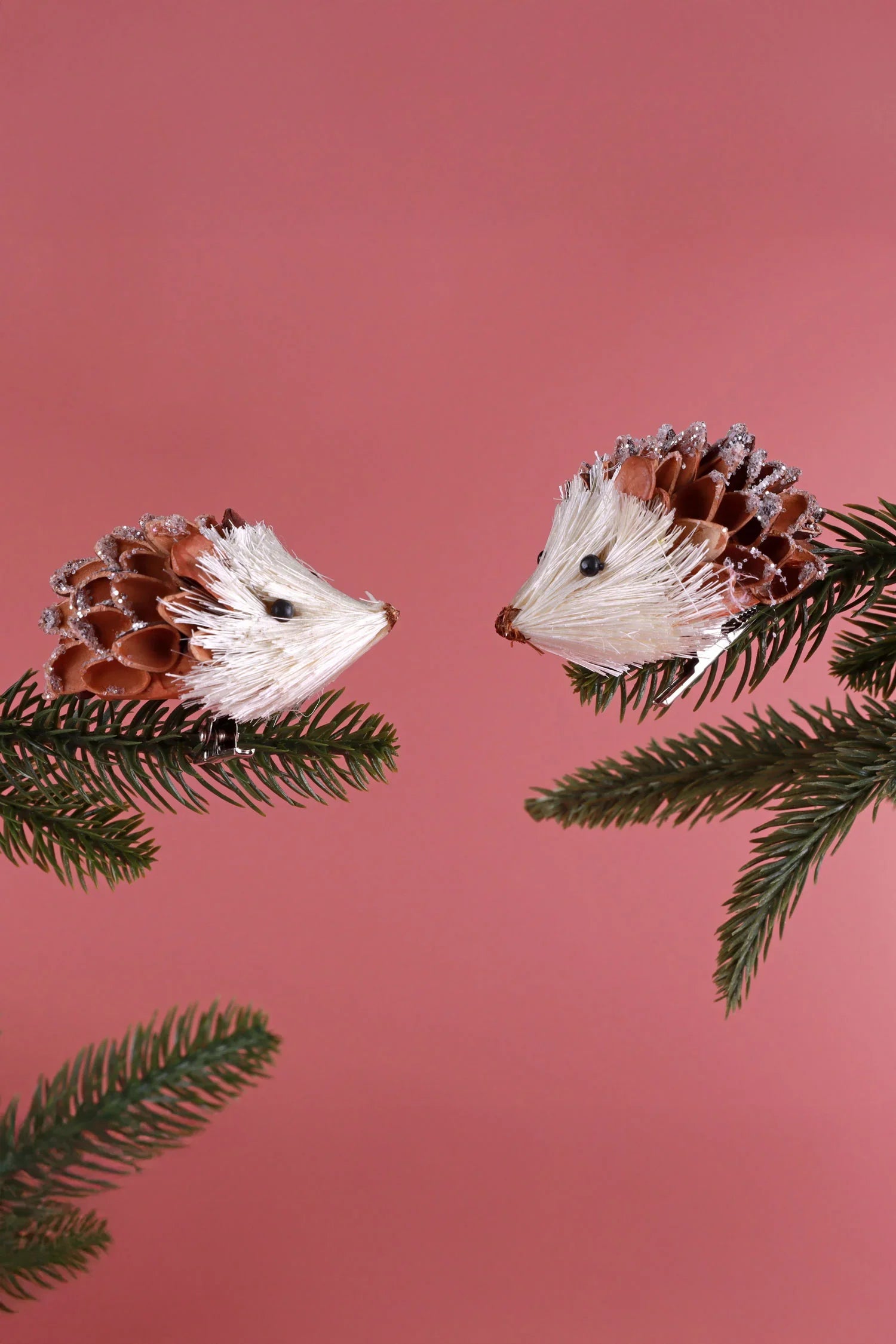 Two pinecone hedgehog ornaments with glitter detail on evergreen branches, pink background