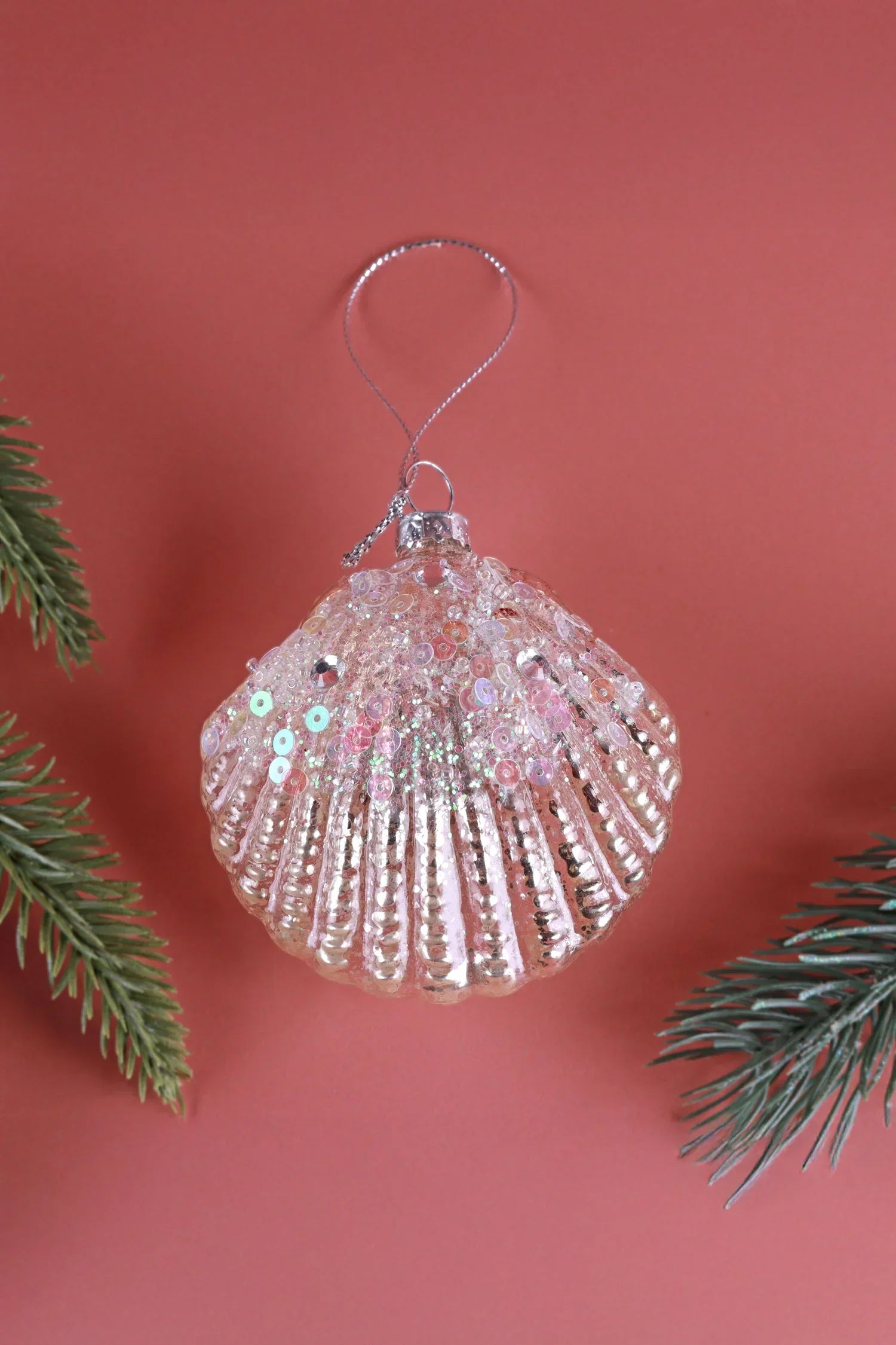 Sequin glass shell ornament by G Decor, hanging against a pink background with pine branches