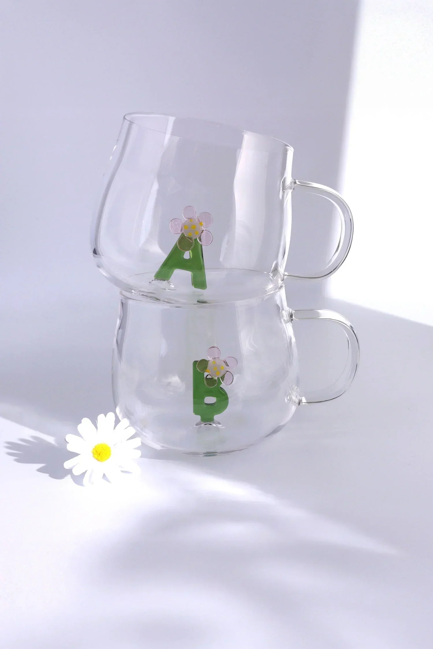 Stacked clear glass mugs with green letter A and B and floral design, daisy on white surface