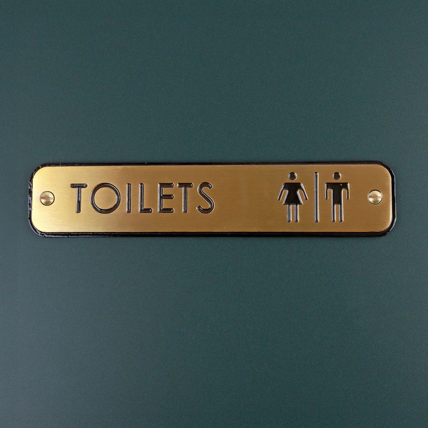 Gold metal toilets sign with male and female icons on a dark green door, G Decor hardware