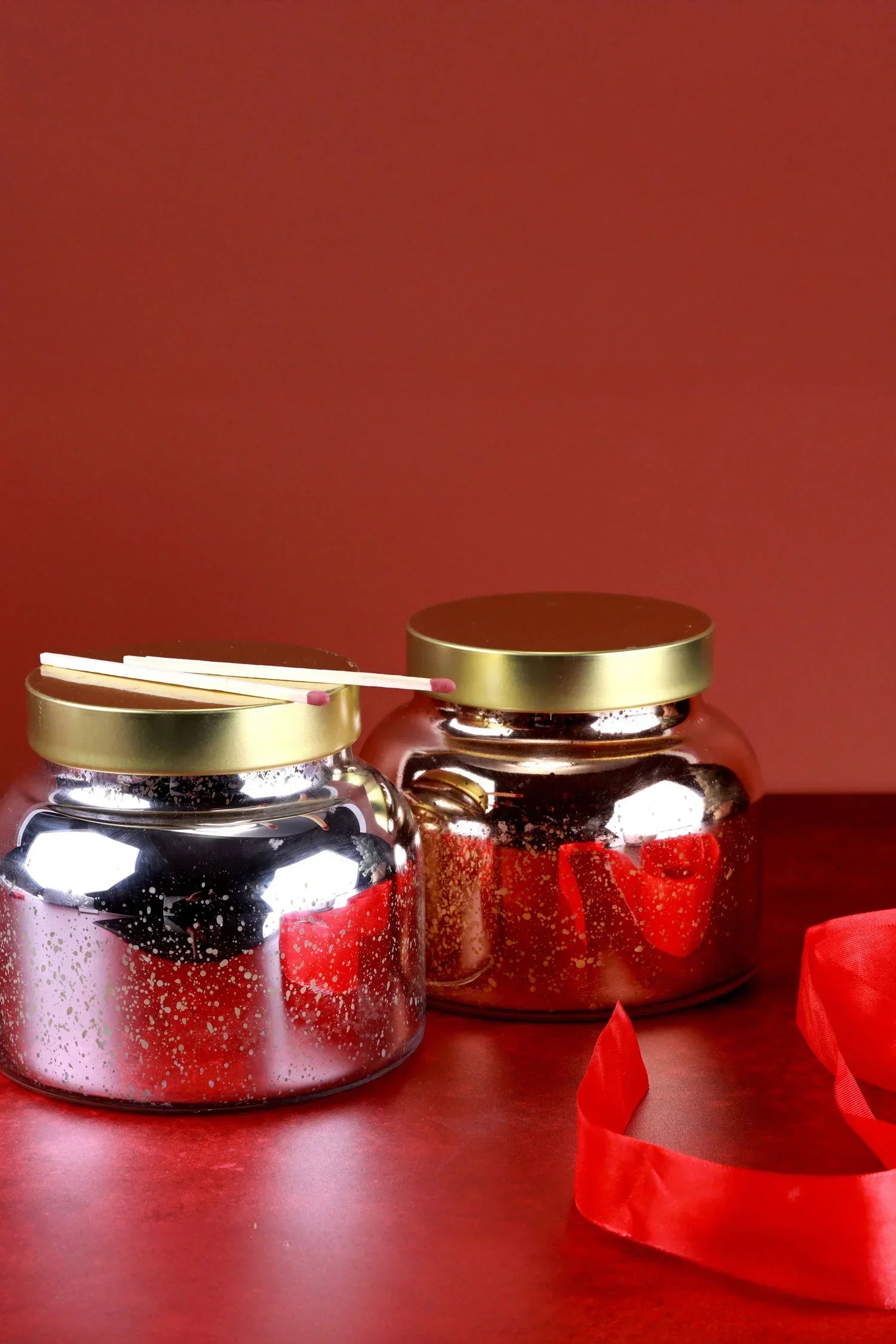 Two metallic glass candles with gold lids and matches, on a red surface by G Decor.