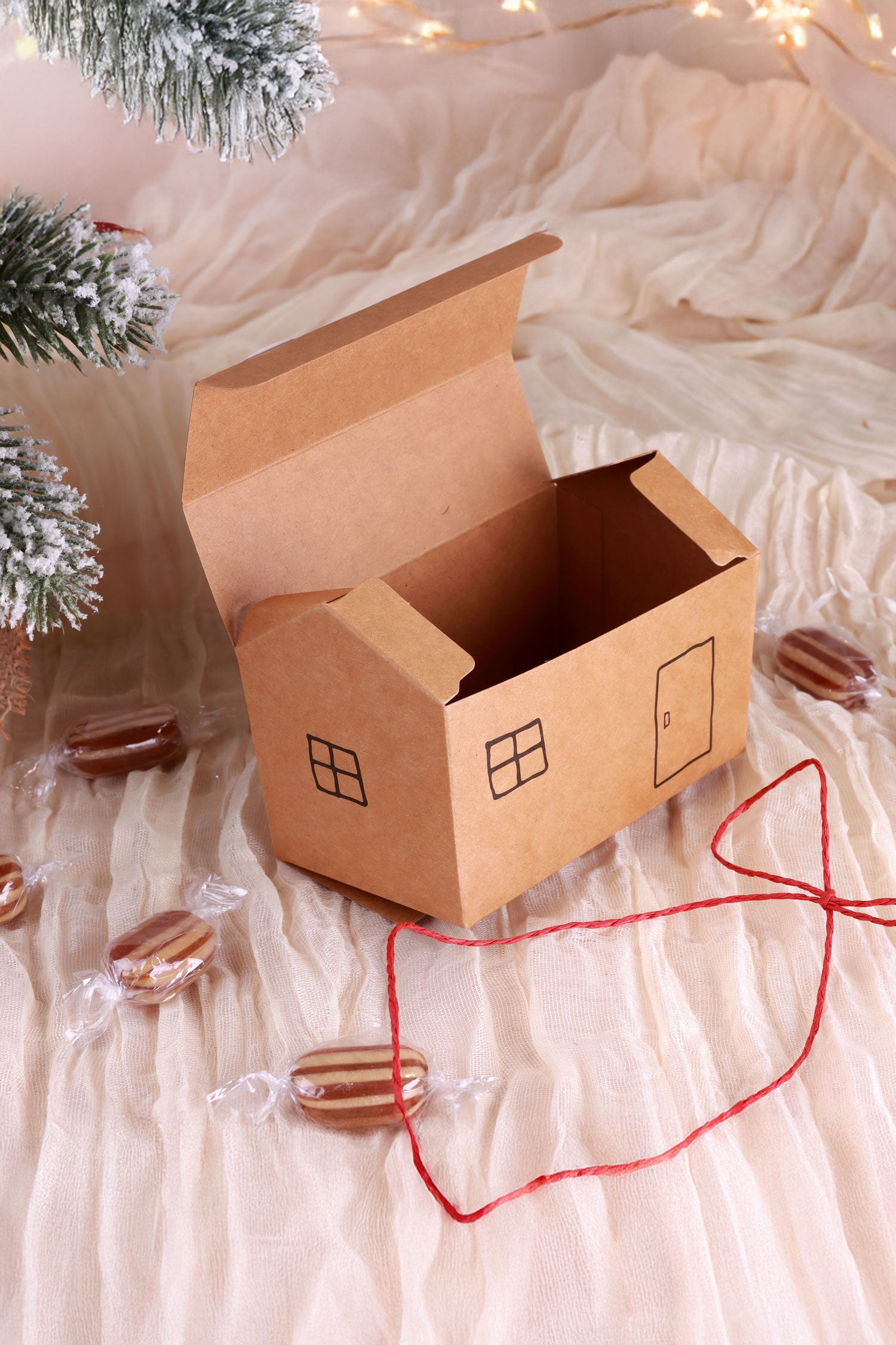 House-shaped kraft paper gift box with window and door print, surrounded by candies and holiday decor