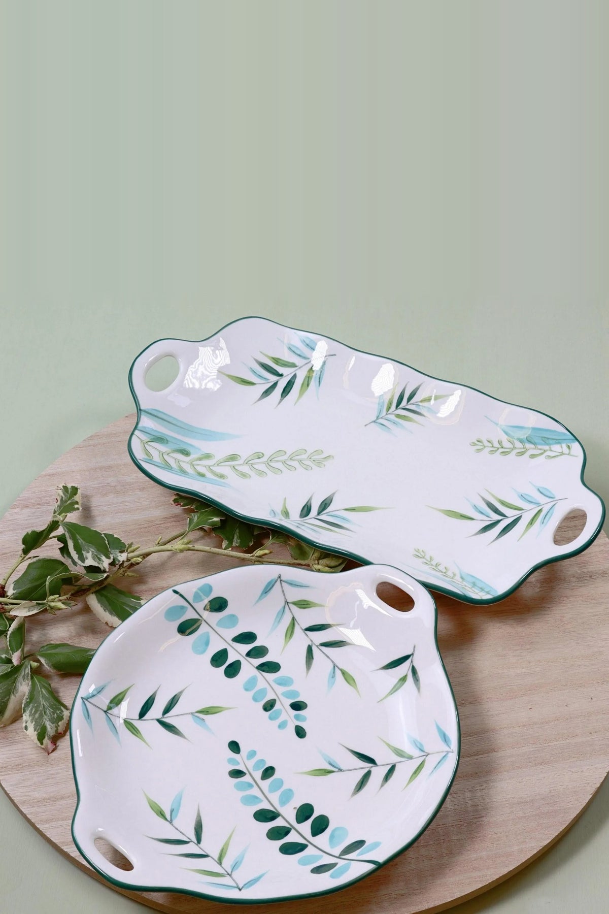 Two ceramic serving plates with green and blue leaf sprays and cut-out handles, a long platter and a round plate, on a wooden board with leaves