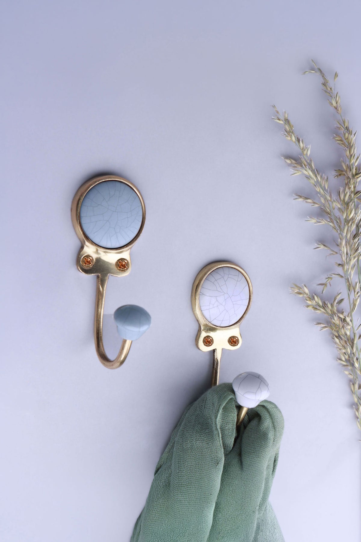 Two crackle glaze ceramic coat hooks, blue-grey and white, on a pale lilac wall with dried grass, a green scarf on the white hook