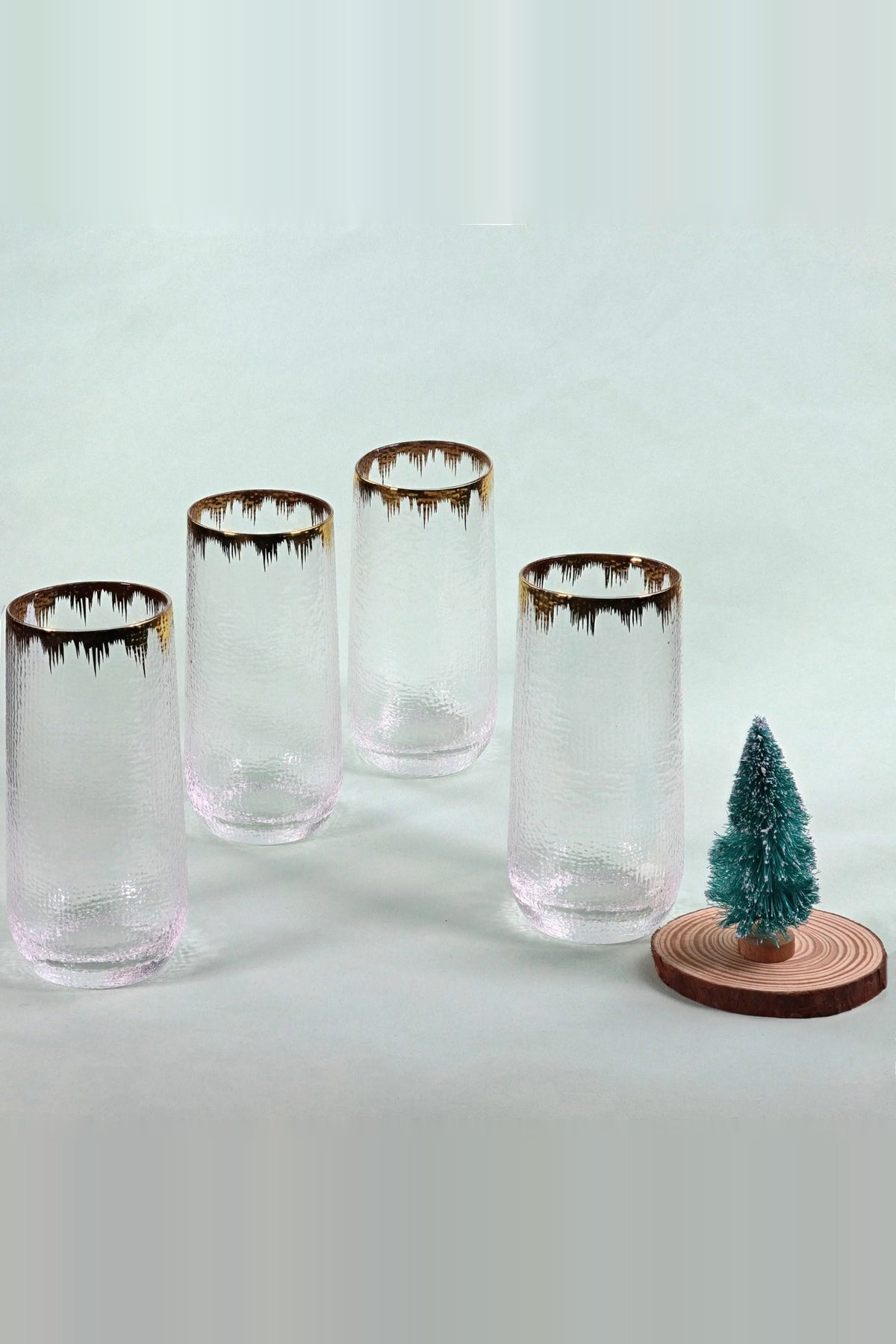 Four tall hammered glass tumblers with gold splash rims on a pale background next to a small Christmas tree on a wood slice