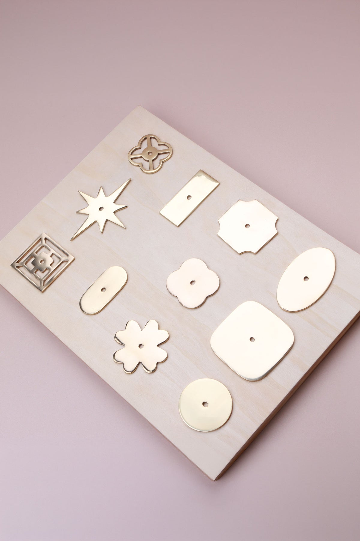 Eleven polished brass cabinet knob backplates on a pale wooden board: round, oval, square, squircle, quatrefoil, pierced, fretwork, flower, star, oblong and rectangle