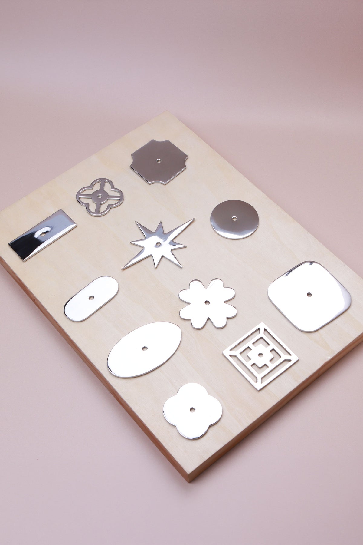 Eleven polished chrome cabinet knob backplates on a pale wooden board: round, oval, square, squircle, quatrefoil, pierced, fretwork, flower, star, oblong and rectangle, second view