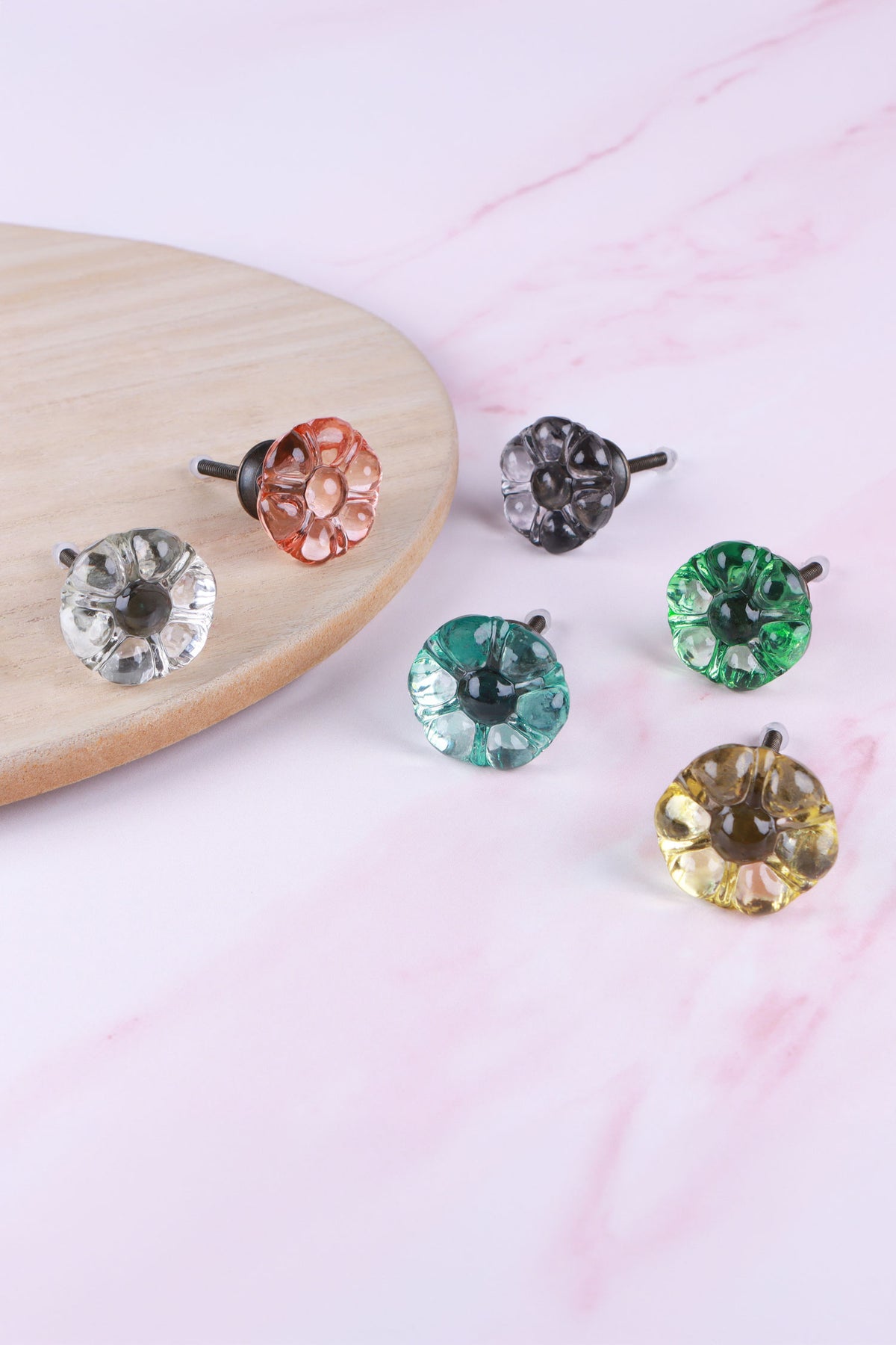 Six-petal flower glass cabinet knobs on antique brass finish bases in clear, blush pink, smokey grey, teal, light green and ivory, on pink marble beside a wooden board