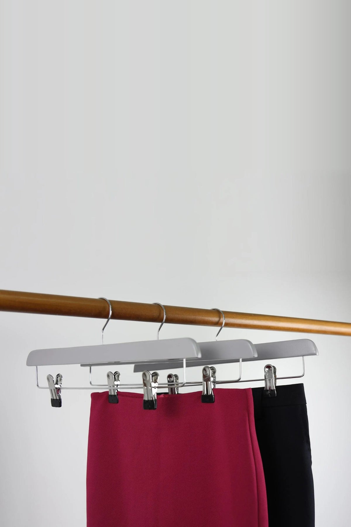 Three grey wooden clip hangers on a wooden rail, holding a red skirt and black trousers