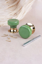 Sage green ceramic door knob standing on its polished brass rose beside the second knob, with the spindle and fixing screws