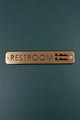 Gold metal restroom sign with modern design on a dark green wall, G Decor luxury hardware.