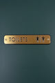 Gold metal toilets sign with male and female icons on a dark green door, G Decor hardware
