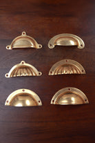 Six aged brass cup pulls — half moon, stepped rim, scalloped, fluted, slim crescent and classic