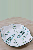 Round white ceramic serving plate with green and blue leaf sprays and two cut-out handles, on a wooden board