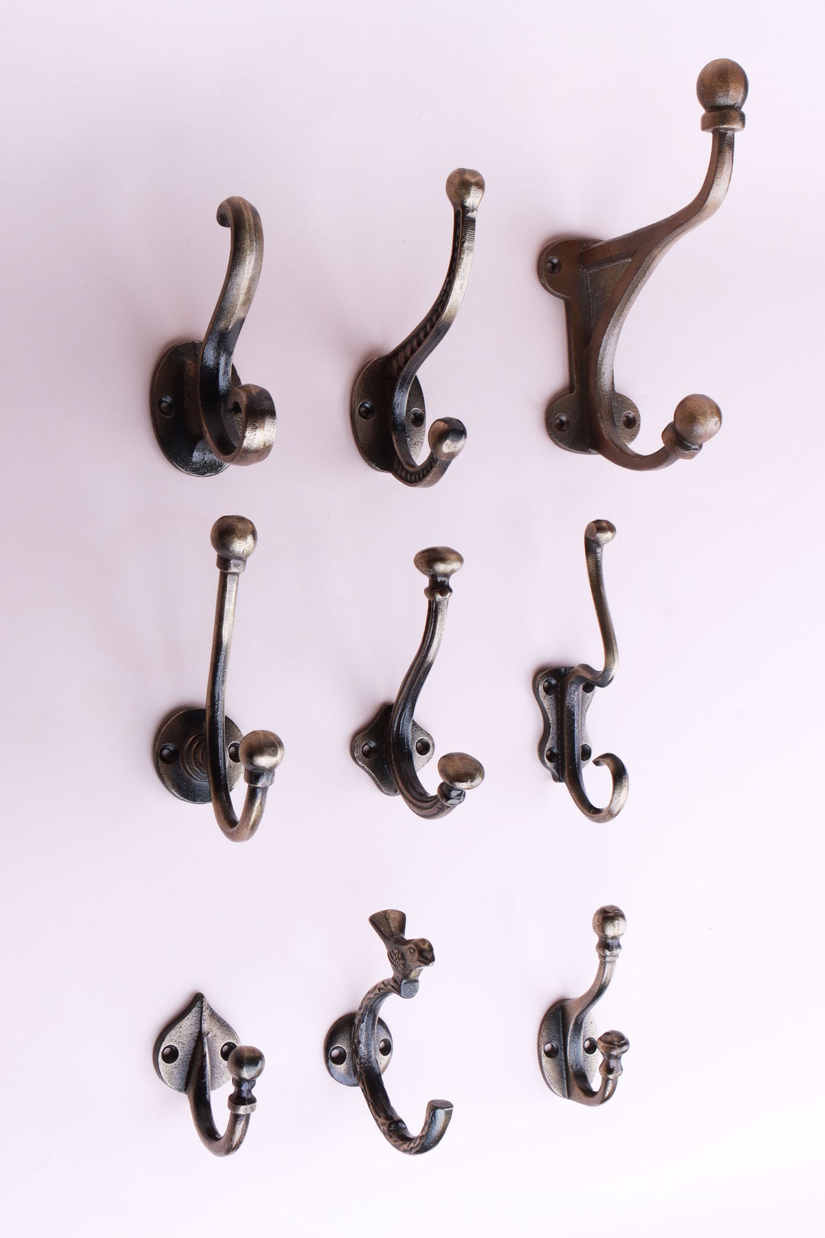 Nine cast iron coat hooks in an antique brass finish on a pale pink wall, from small single hooks to a large hat and coat hook