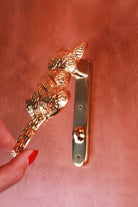 Hand lifting the brass cat door knocker by its tail, showing the slim back plate, its two screws and the round strike button