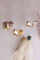 Four ceramic ball hooks with a brass finish on a pale pink wall with a sprig of white buds: rosy brown, cream, mustard yellow and green, a cream scarf on the green hook