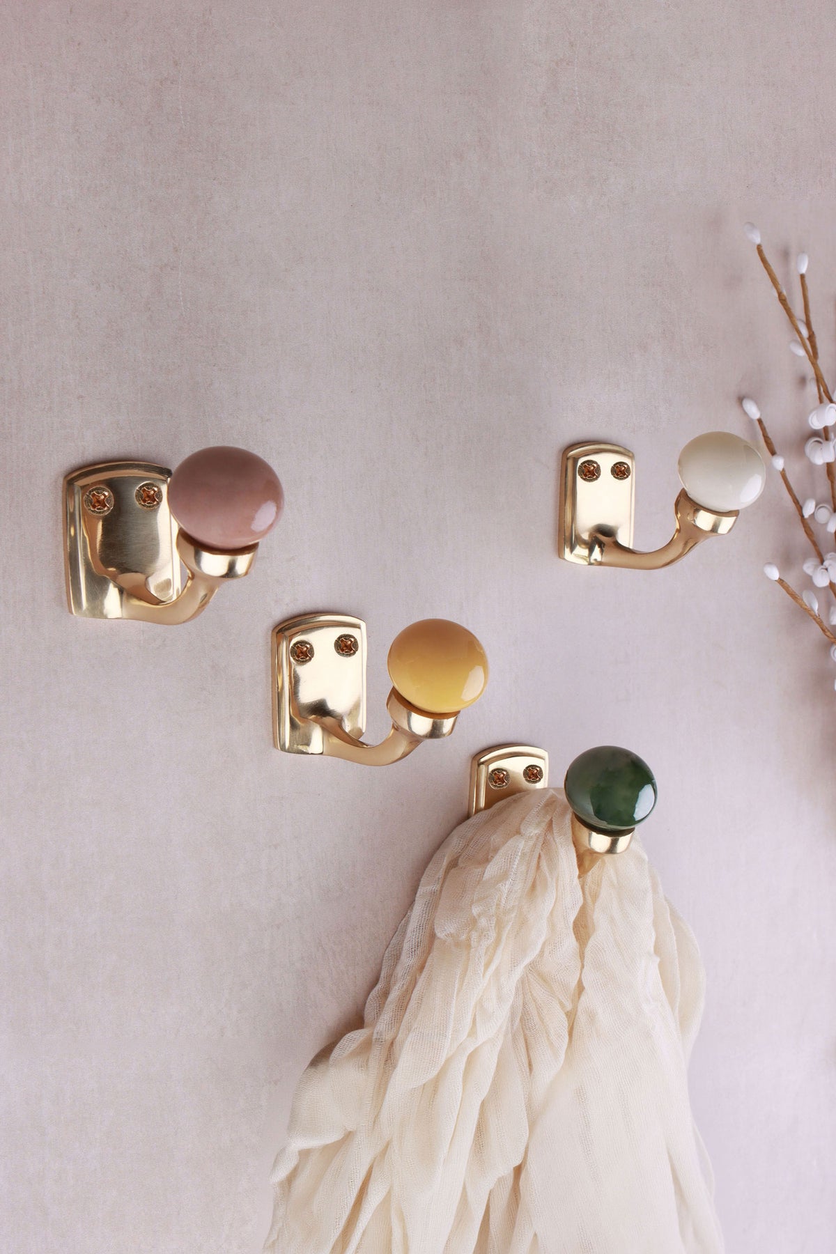 Four ceramic ball hooks with a brass finish on a pale pink wall with a sprig of white buds: rosy brown, cream, mustard yellow and green, a cream scarf on the green hook