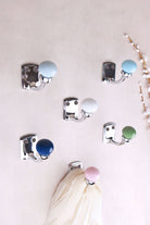 Six chrome finish ceramic ball hooks on a pale wall with white buds: light grey, light blue, white, navy blue, sage green and light pink, a cream scarf on the pink hook