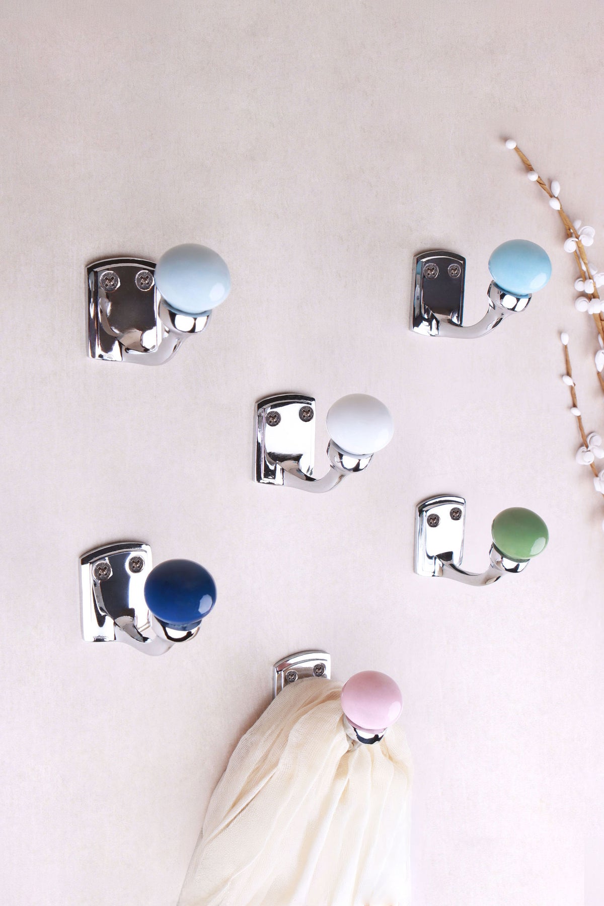 Six chrome finish ceramic ball hooks on a pale wall with white buds: light grey, light blue, white, navy blue, sage green and light pink, a cream scarf on the pink hook