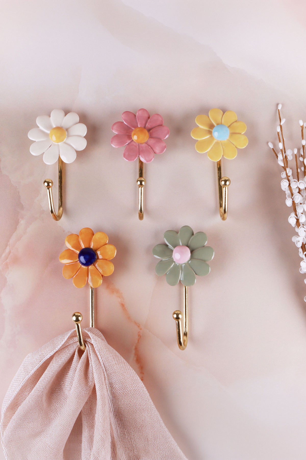 Five ceramic daisy wall hooks in white, pink, yellow, dark yellow and sage green, with a scarf hanging from one