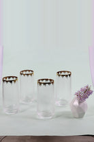 Four hammered glass tumblers with gold rims next to a white vase of lilac flowers