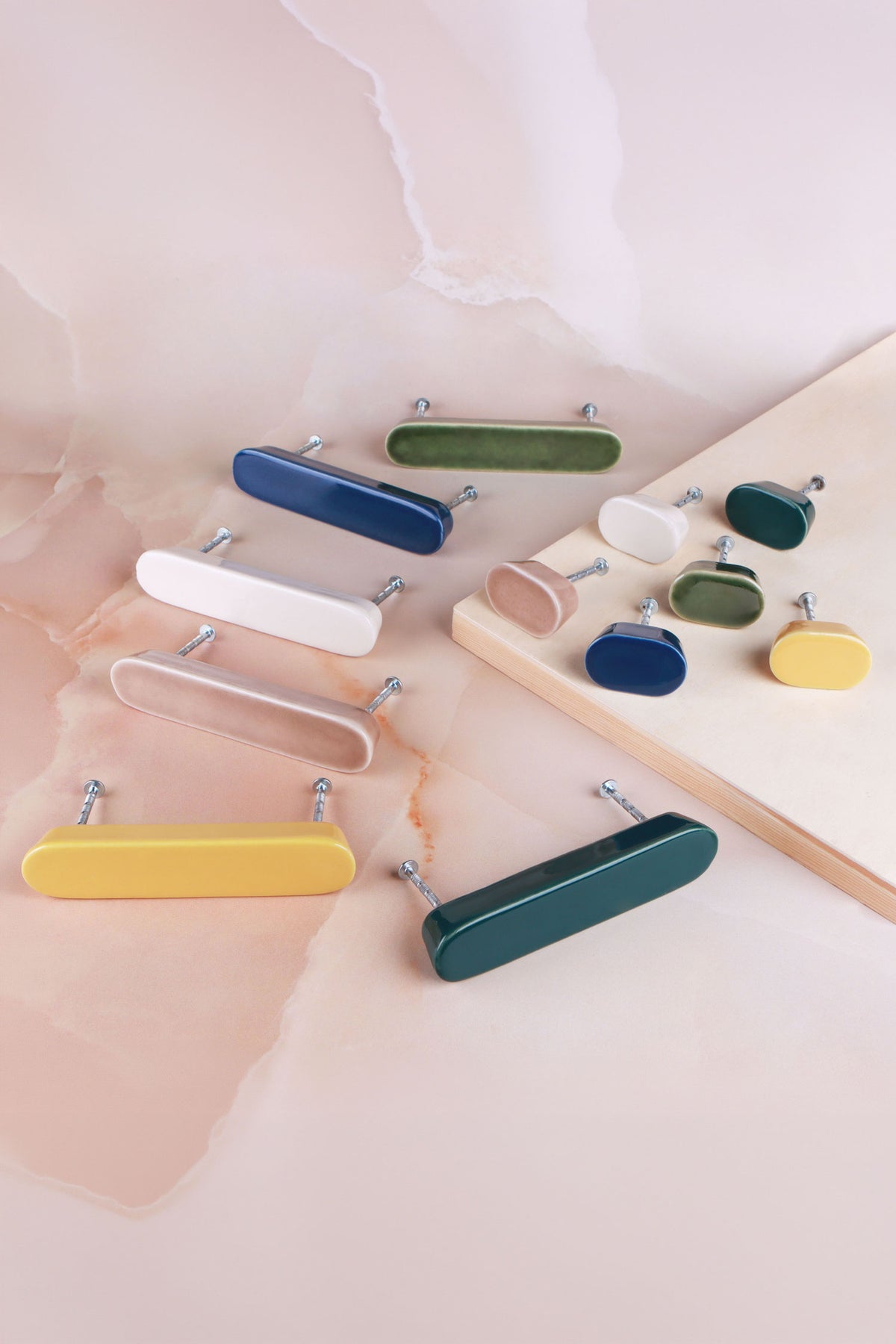Oval ceramic cabinet knobs and matching bar handles in cream, brown, yellow, olive green, dark teal and cobalt blue on pink marble with a wooden board