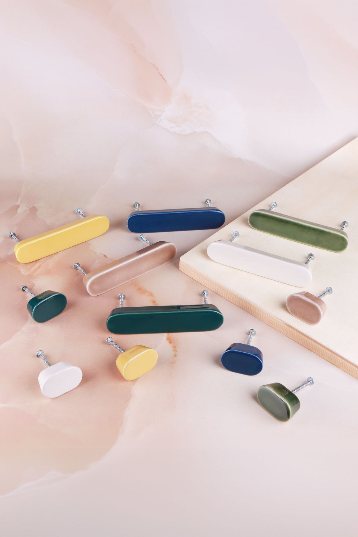 Six oval ceramic knobs and six long ceramic handles laid out on pink marble and a wooden board