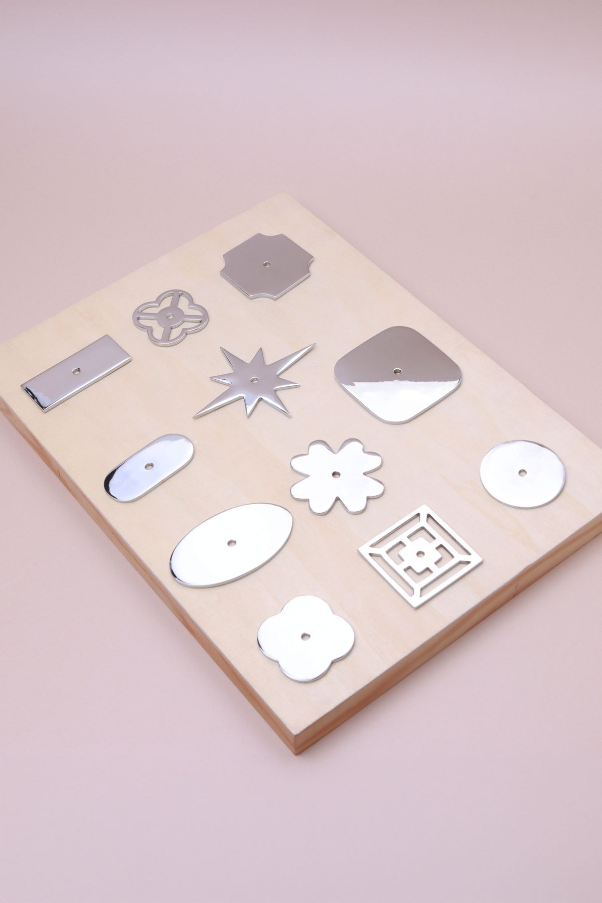 Eleven polished chrome cabinet knob backplates on a pale wooden board: round, oval, square, squircle, quatrefoil, pierced, fretwork, flower, star, oblong and rectangle