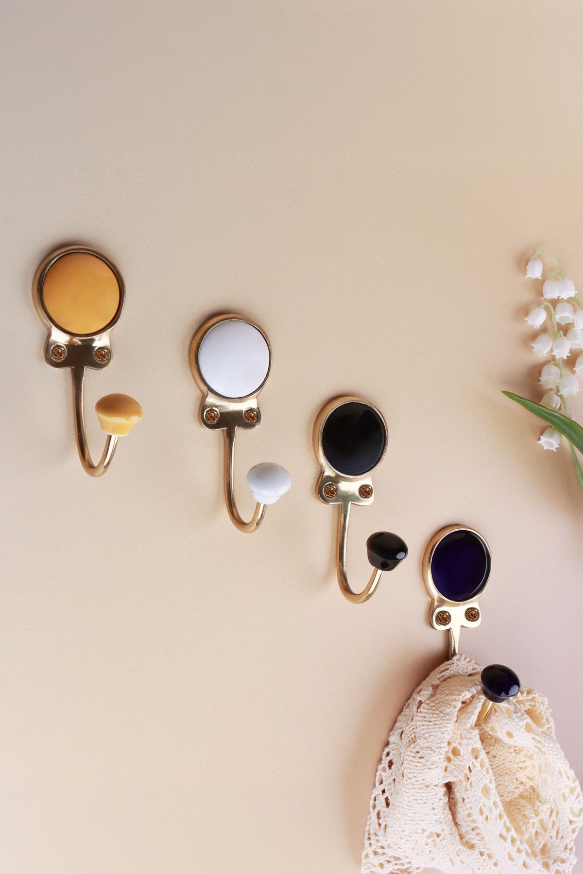 Four ceramic coat hooks in yellow, white, black and dark blue on a peach wall with lily of the valley, a lace cloth on the dark blue hook