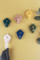 Six scallop shell ceramic wall hooks on a warm yellow wall with eucalyptus: dark teal, brown, yellow, olive green, cream and cobalt blue, a cloth on the cream hook